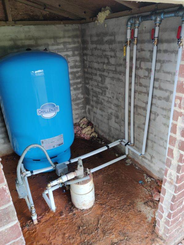 A blue water pressure tank with white pipes connected in a small, unfinished brick room. Several valves are attached to the pipes, and the floor is bare concrete.