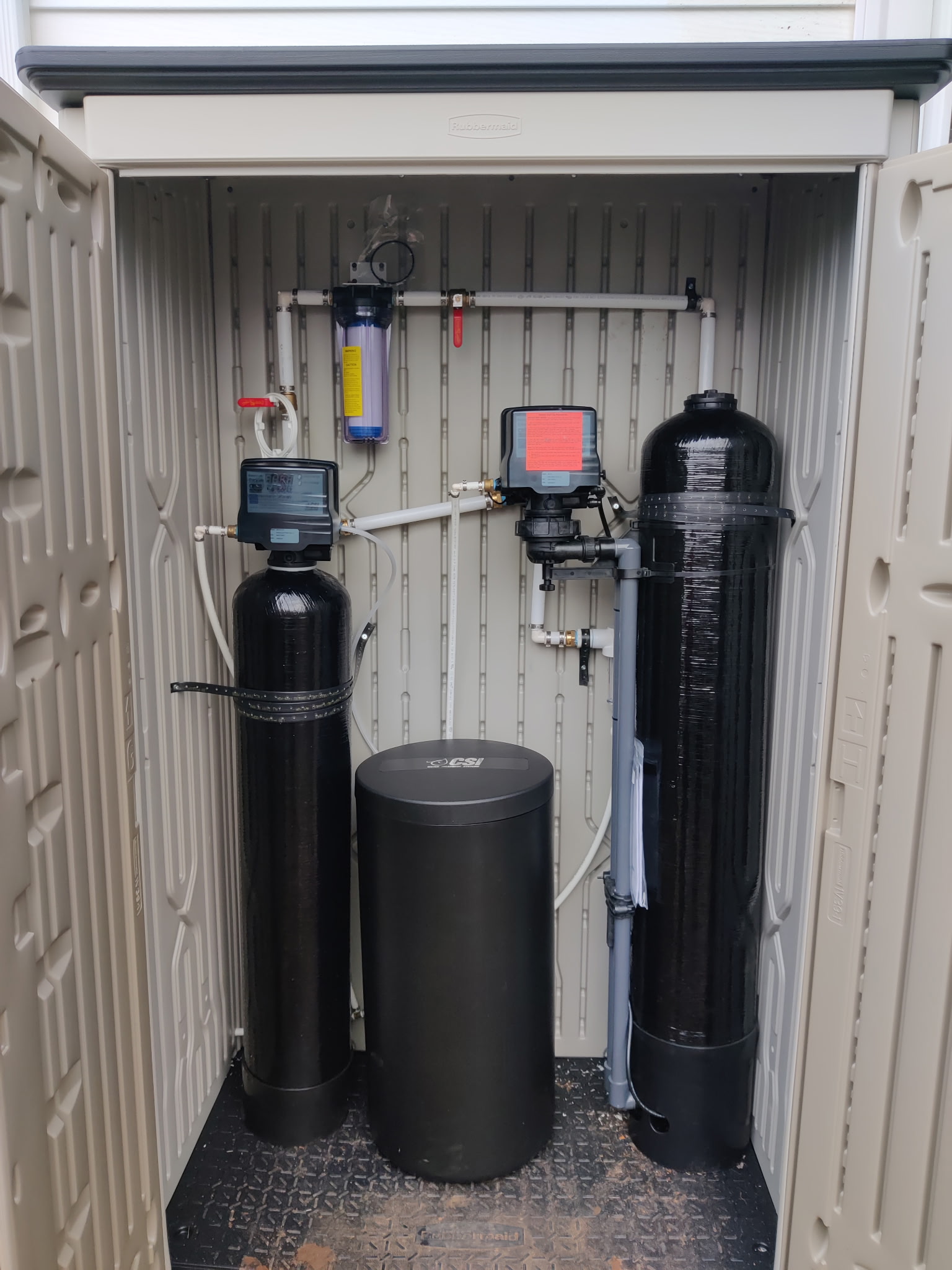 Water filtration system inside a plastic enclosure, featuring two tall black tanks, a central cylindrical unit, and visible pipes, valves, and a gauge.