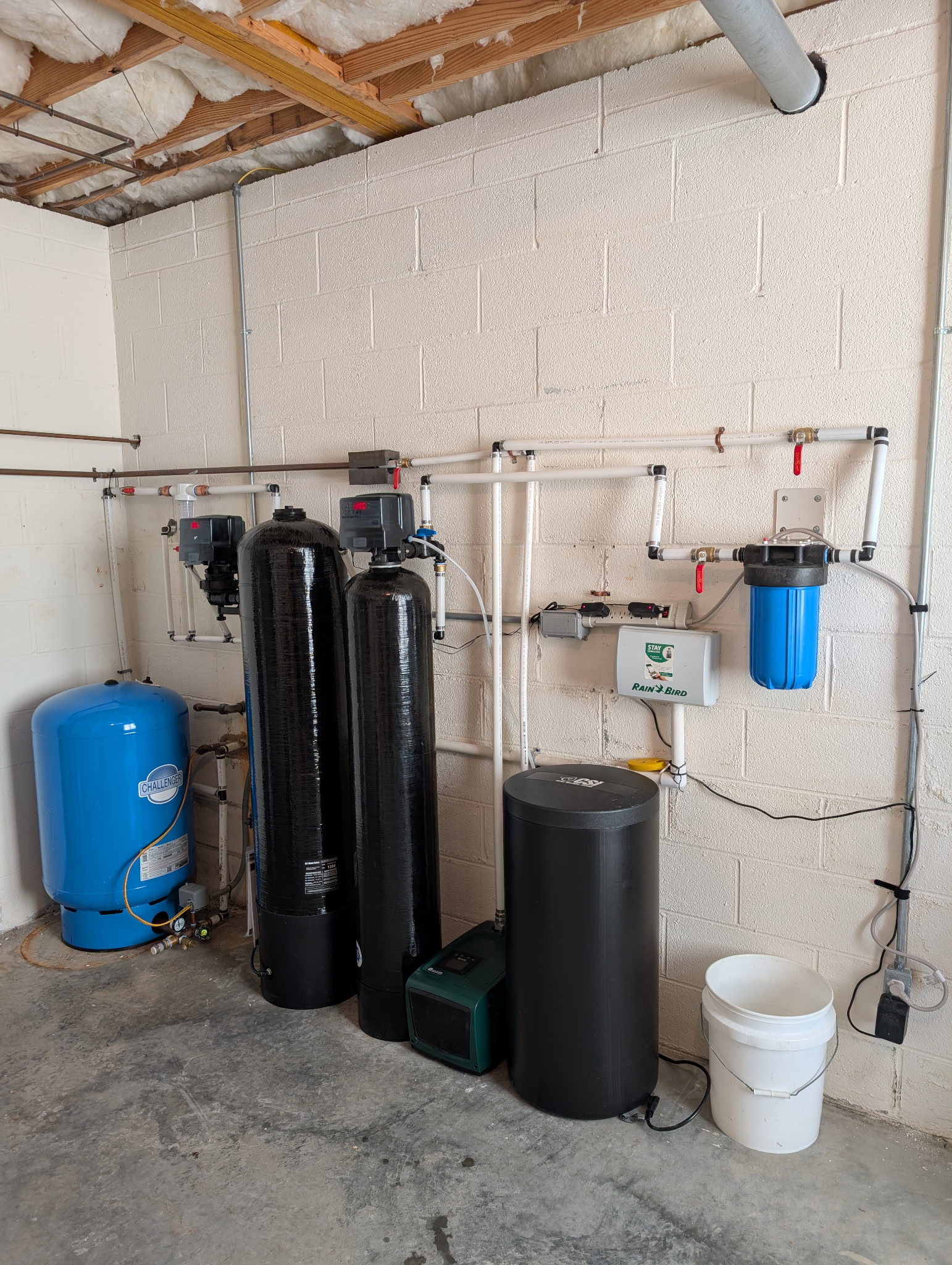 Water filtration system setup with blue pressure tank, two black filter units, a blue filter, control boxes, pipes, and a white bucket on a concrete floor against a white brick wall.