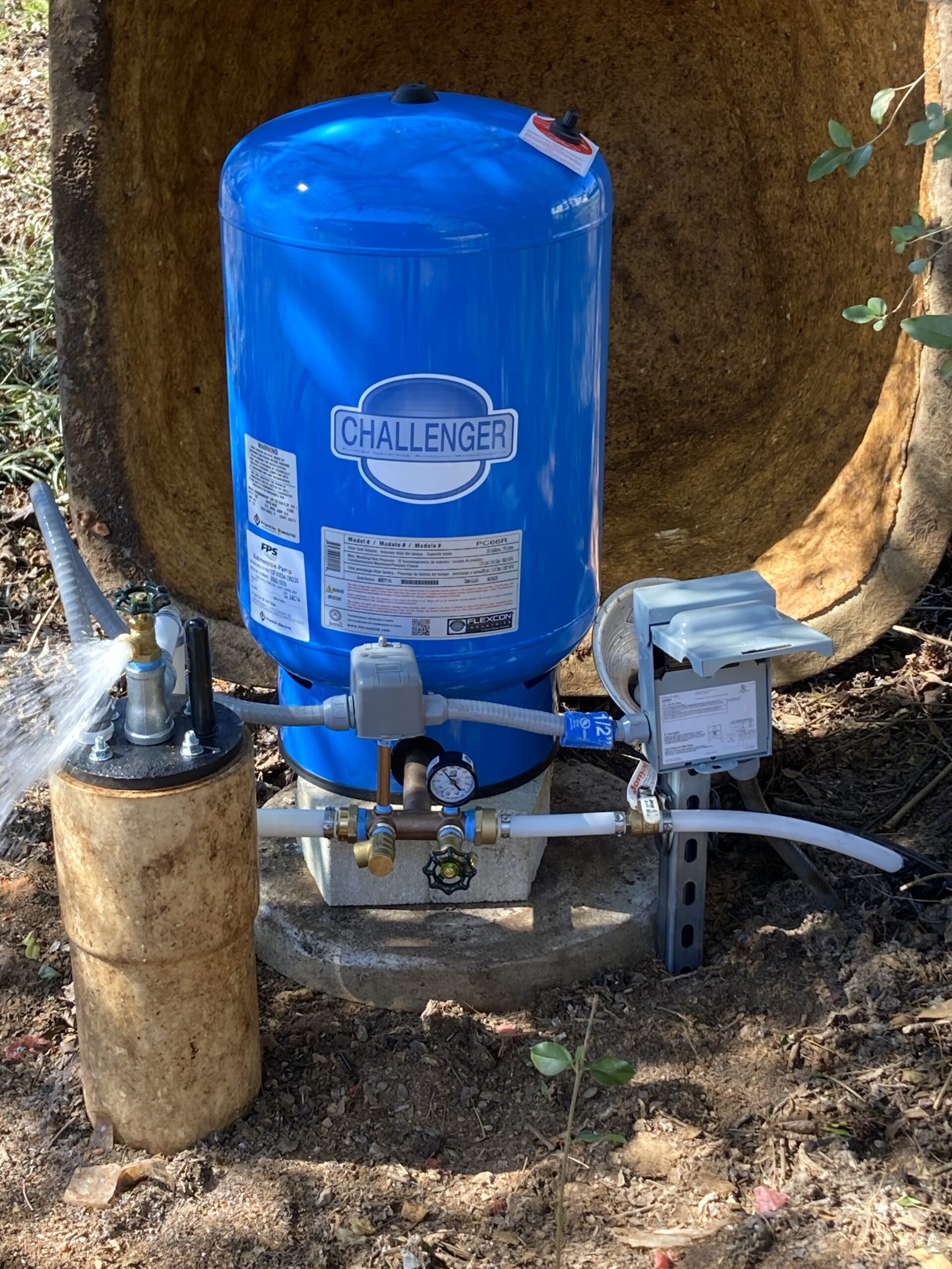 A blue Challenger pressure tank with attached pipes and a gauge is installed outdoors on a concrete base, surrounded by dirt and vegetation.